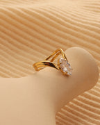 Anillo abierto dorado de bandas curvas con piedra transparente de forma alargada, sobre soporte beige.