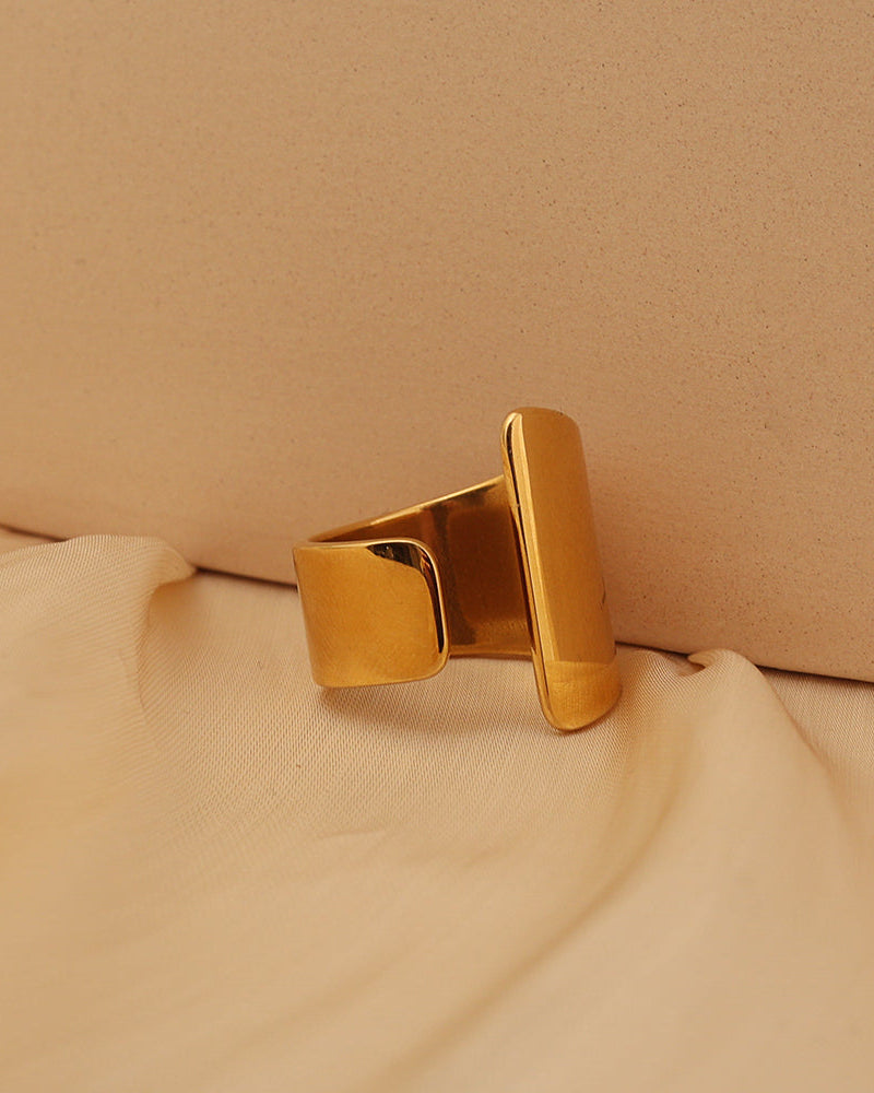 Anillo abierto dorado con dos extremos rectangulares lisos de diferente longitud, sobre tela beige.