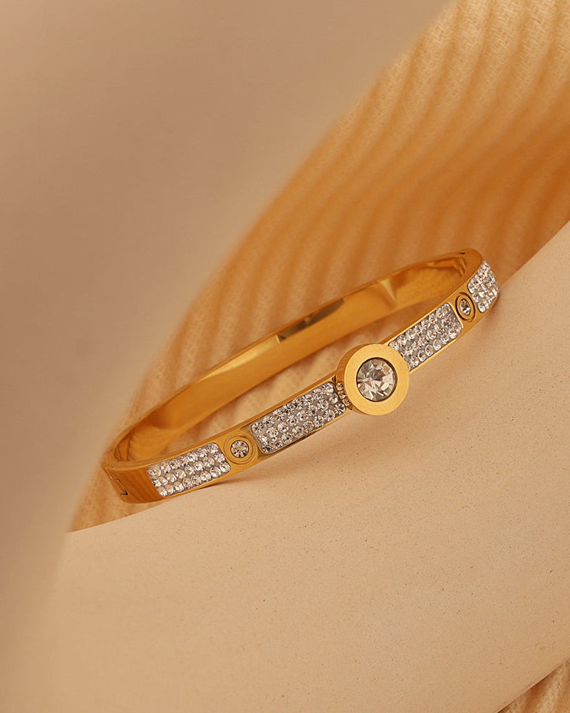 Pulsera rígida dorada con motivos circulares y tramos de pequeñas piedras claras, sobre soporte beige.