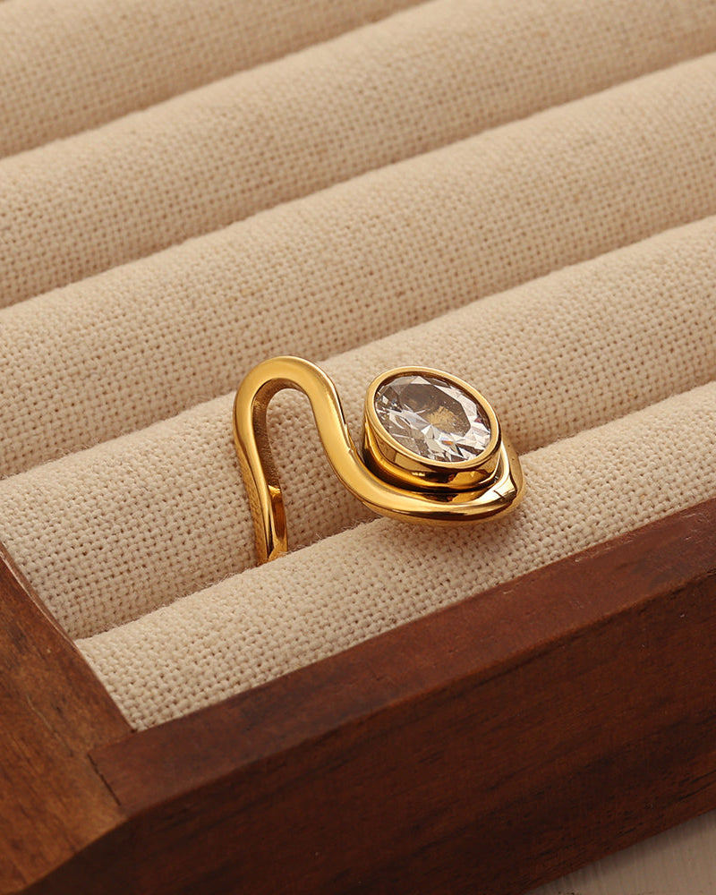 Anillo abierto dorado de banda curva con piedra transparente ovalada, en un joyero de tela beige.