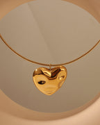 Collar dorado con corazón grande abombado de superficie irregular y brillante, sobre fondo crema.