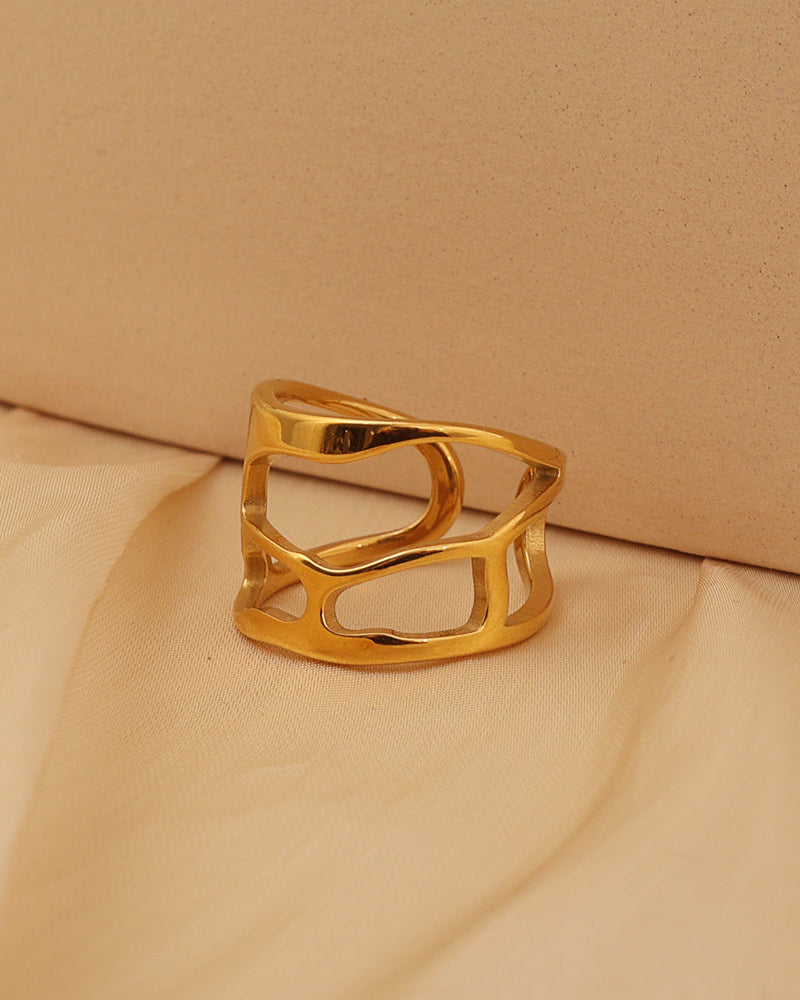 Anillo ancho dorado calado con formas curvas entrecruzadas, sobre tela beige.