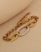 Pulsera dorada de cadena con grandes eslabones ovalados centrales, uno cubierto de pequeñas piedras claras, sobre tela beige.