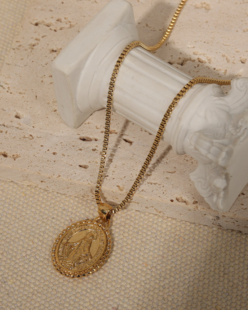 Collar dorado con medallón ovalado de borde ornamentado, dispuesto junto a una columna blanca sobre piedra y tela beige.