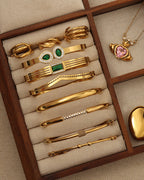 Joyero con varias pulseras rígidas doradas, algunas con piedras verdes o detalles entrelazados, y un collar rosa.