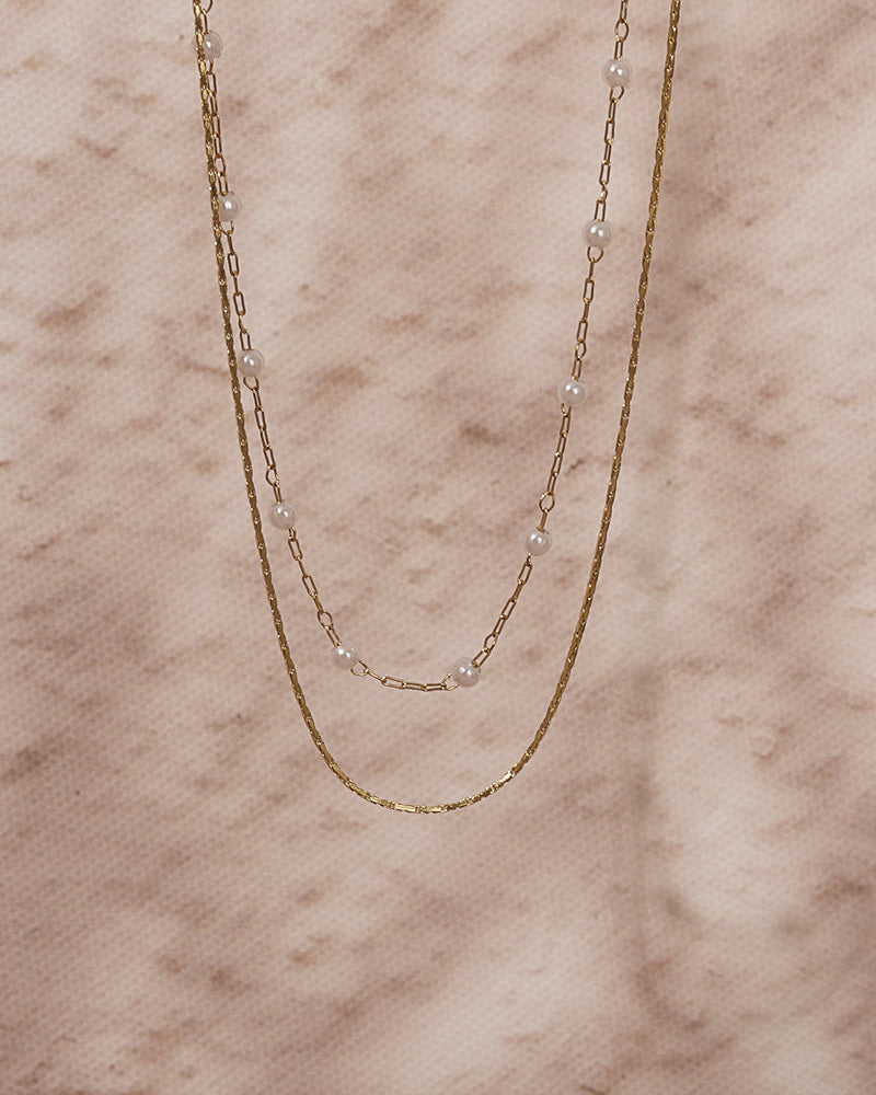 Dos collares finos dorados, uno con pequeñas perlas blancas, extendidos sobre mármol rosado.