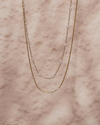Dos collares finos dorados, uno con pequeñas perlas blancas, extendidos sobre mármol rosado.