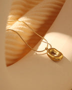 Collar fino dorado con colgante ovalado irregular de superficie hundida y pulida, sobre fondo beige al sol.