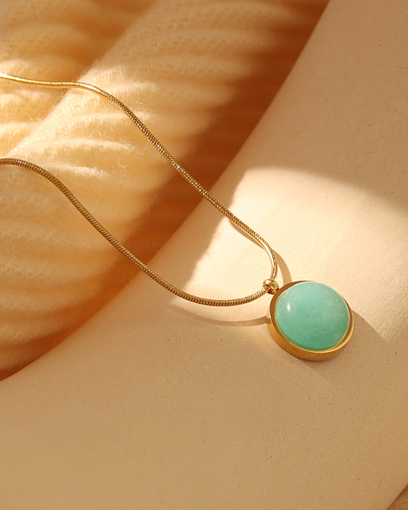 Collar dorado con colgante redondo verde turquesa sobre fondo beige iluminado por el sol.