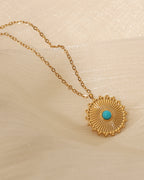 Colgante circular dorado con rayos y pequeño centro azul turquesa, sobre tela clara translúcida.