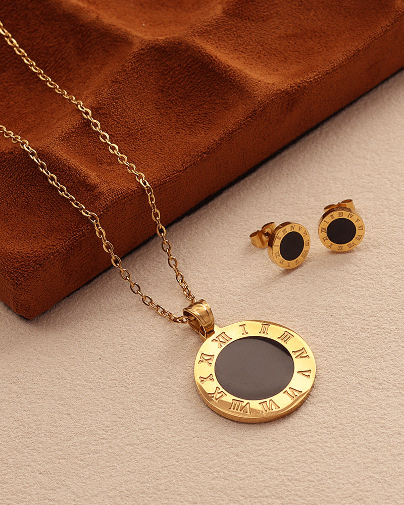 Collar de medallón negro con borde dorado de números romanos y pendientes a juego, junto a estuche marrón sobre tela clara.