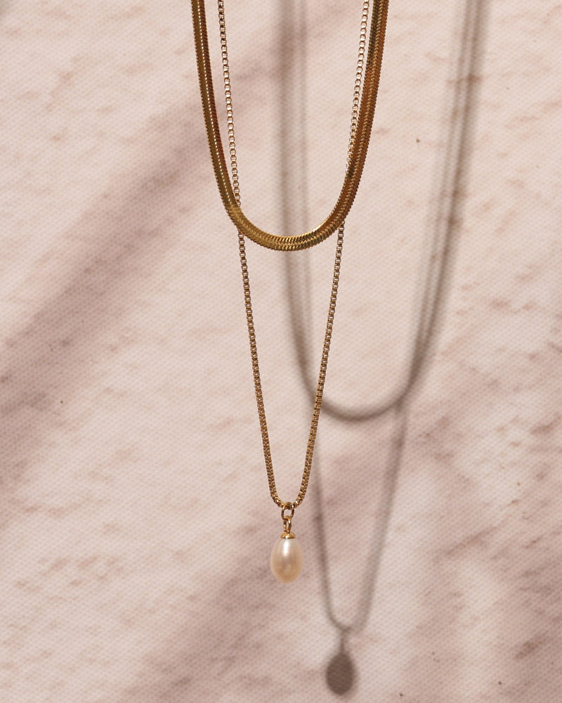 Collar doble dorado de cadena plana y cadena fina con perla blanca, colgado sobre mármol rosado.