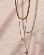 Collar doble dorado de cadena plana y cadena fina con perla blanca, colgado sobre mármol rosado.