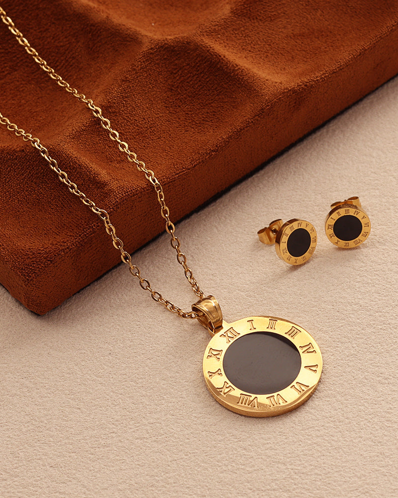 Collar dorado de medallón negro con borde de números romanos y pendientes a juego, junto a un estuche marrón.