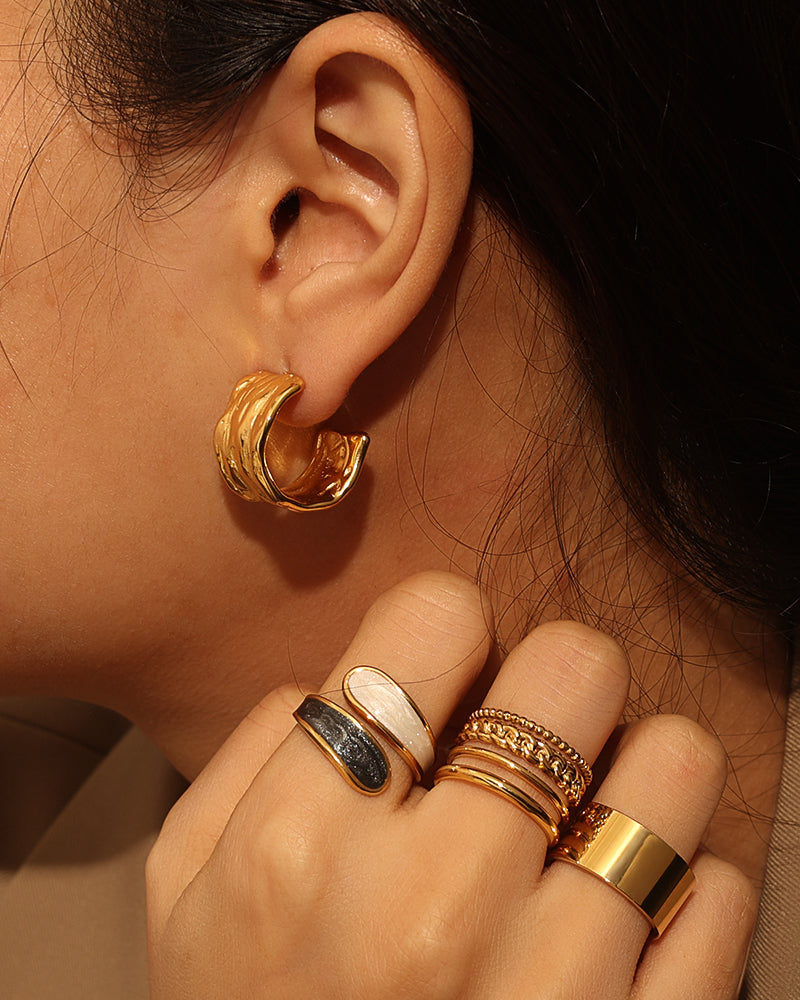 Persona con pendiente de aro grueso dorado de varias bandas curvas y varios anillos en la mano.