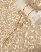 Collar dorado tipo rosario con medalla y cruz, dispuesto junto a una pequeña columna blanca con reflejos de luz.