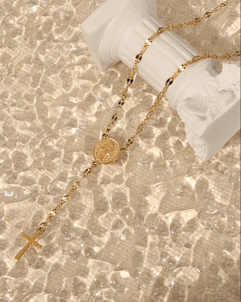 Collar dorado tipo rosario con medalla y cruz, dispuesto junto a una pequeña columna blanca con reflejos de luz.