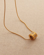 Collar fino dorado con colgante cilíndrico y pequeños motivos de piedras claras, sobre tela beige.