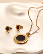 Collar dorado de medallón negro con números romanos y pendientes a juego, sobre superficie satinada clara.