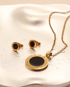Pendientes redondos dorados de centro negro junto a collar de medallón a juego, sobre superficie satinada clara.