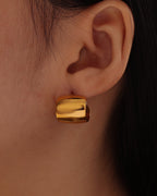 Pendiente dorado ancho, curvo y pulido de forma rectangular redondeada, puesto en la oreja.