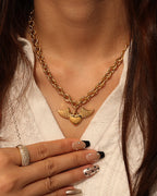 Persona con collar de cadena dorada y colgante de corazón con alas, sobre blusa blanca.