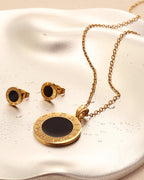 Conjunto de collar dorado con medallón negro y pendientes redondos a juego, sobre superficie satinada clara.
