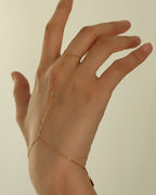 Pulsera fina dorada unida por una cadena a un anillo, puesta en la mano extendida.