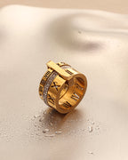 Anillo ancho dorado de varias bandas, números romanos y pequeñas piedras claras, sobre superficie satinada clara.
