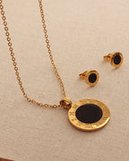 Conjunto de collar de cadena dorada y pendientes de discos negros con bordes dorados grabados, sobre tela beige.