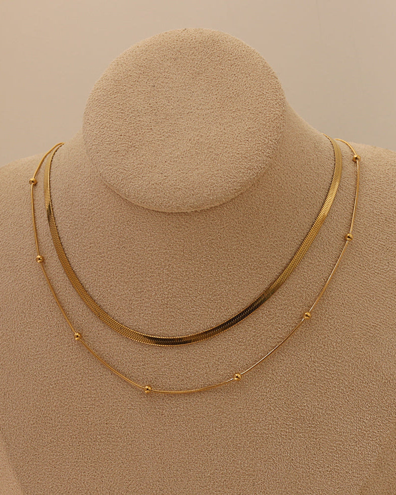Dos cadenas doradas, una plana y otra fina con pequeñas esferas, sobre un busto de tela beige.