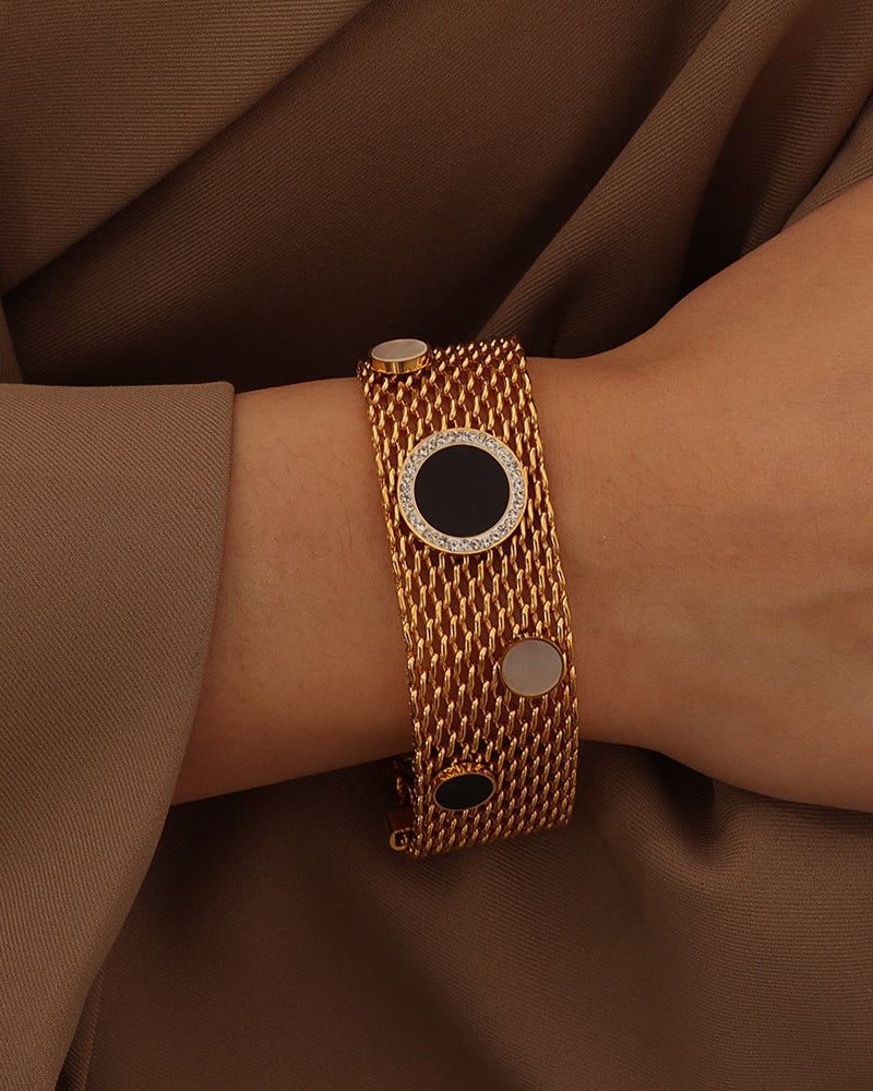 Pulsera ancha de malla dorada con círculos negros y claros, puesta en la muñeca sobre tela marrón.