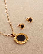 Collar de medallón circular negro con borde dorado de números romanos y pendientes a juego, sobre tela beige.