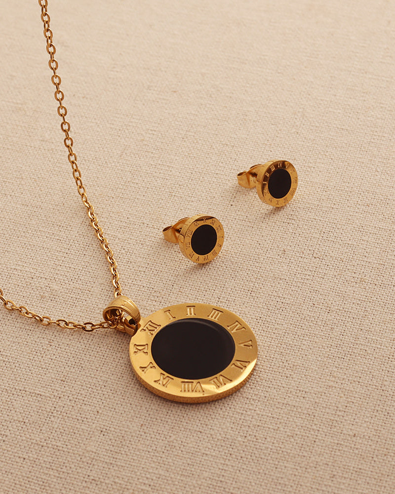 Collar de medallón circular negro con borde dorado de números romanos y pendientes a juego, sobre tela beige.