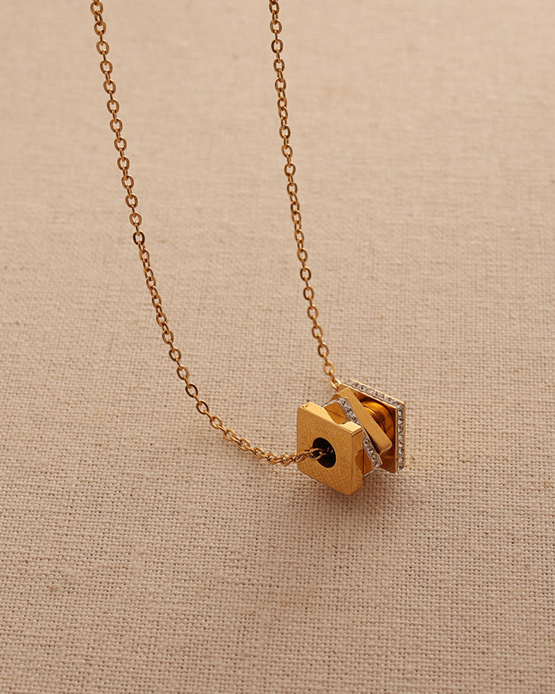 Collar dorado de cadena fina con colgante geométrico de bloques dorados y detalles oscuros, sobre tela beige.