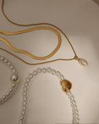 Collar doble de cadena plana dorada y cadena fina con perla colgante, junto a una pulsera de perlas y corazón dorado.