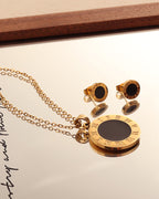 Collar dorado de medallón negro y pendientes redondos a juego, con su reflejo sobre espejo de marco de madera.