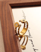 Anillo abierto dorado con discos pequeños en los extremos, uno negro y otro claro, reflejado en un espejo con marco de madera.