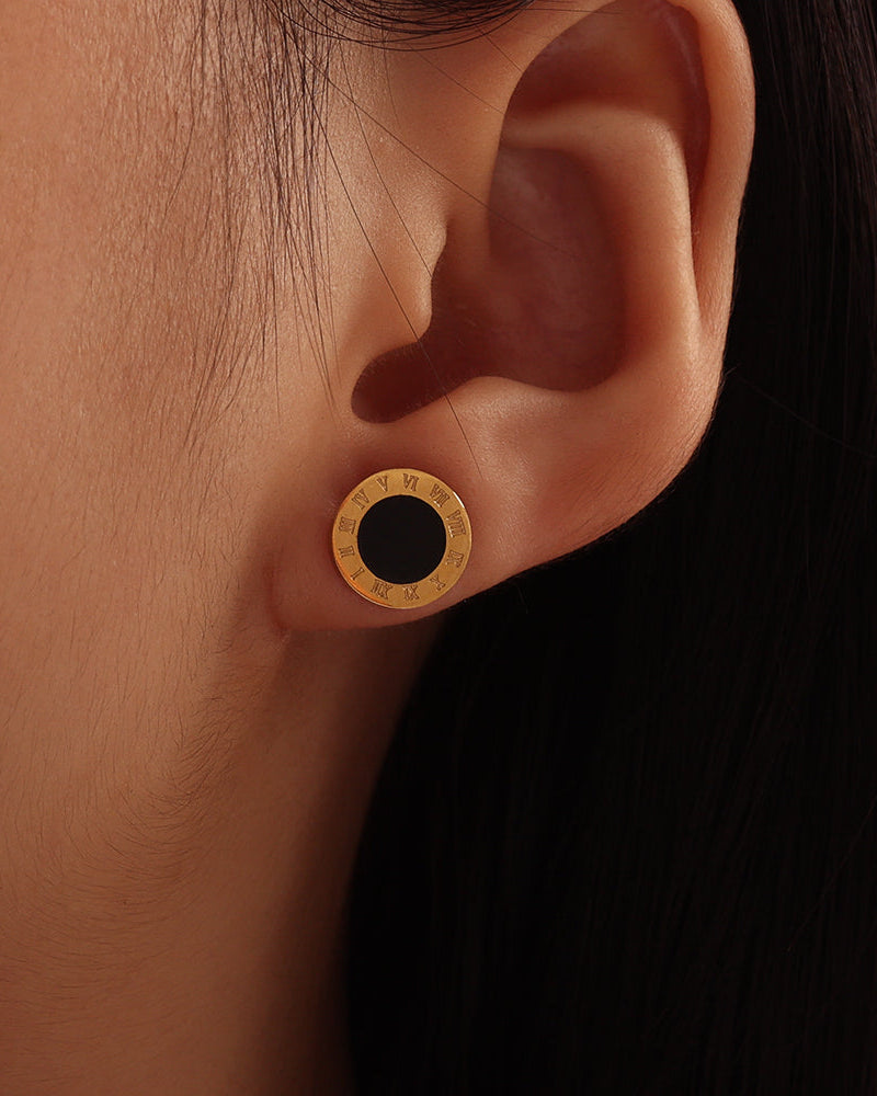 Pendiente redondo de centro negro y borde dorado grabado, puesto en la oreja junto al cabello oscuro.