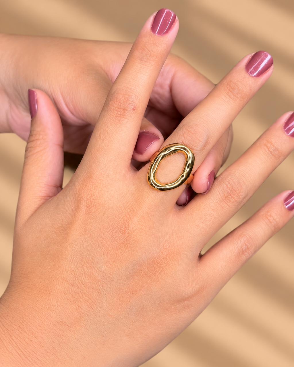 Anillo dorado de gran óvalo abierto con borde brillante, puesto en una mano con uñas moradas.