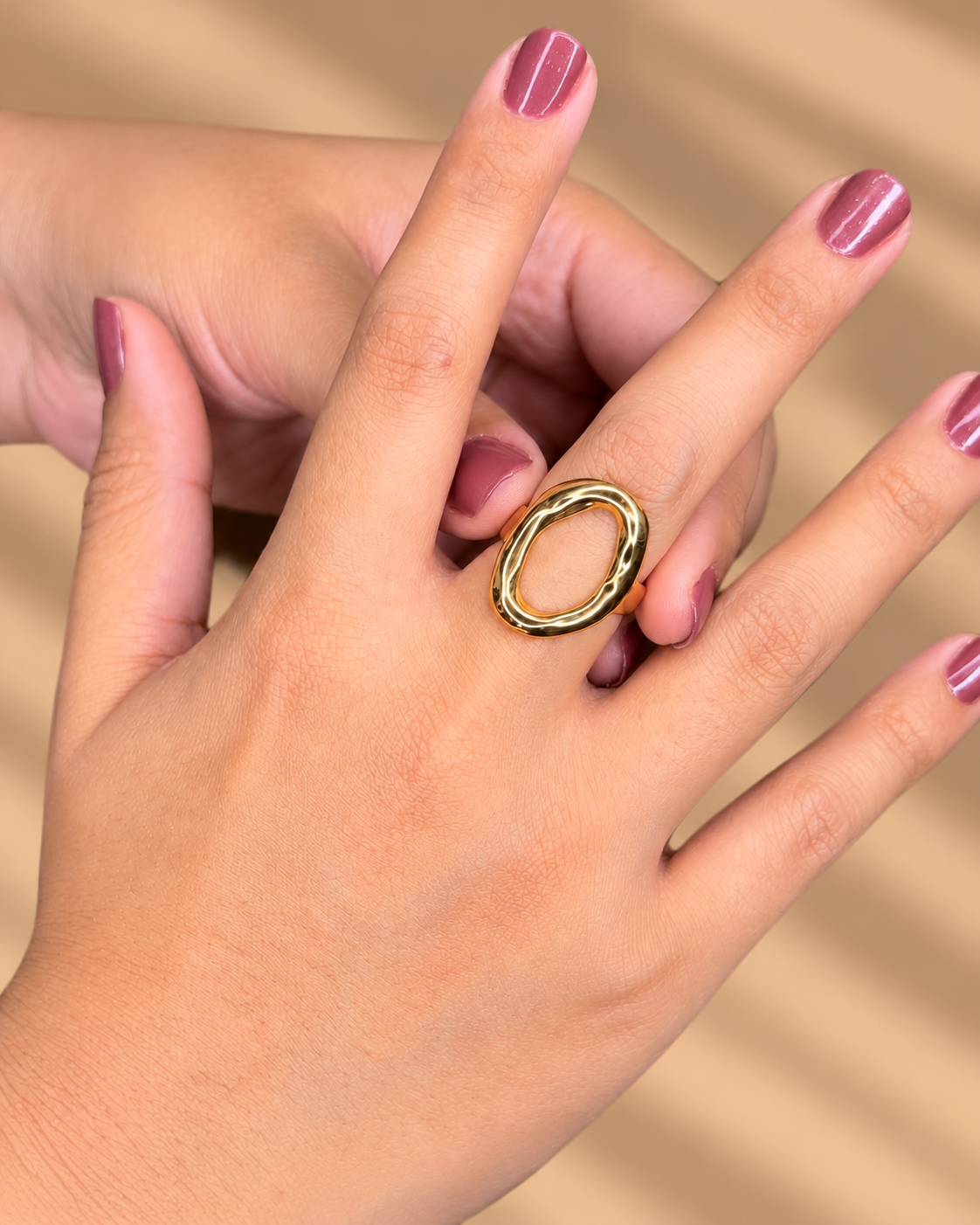 Anillo dorado de gran óvalo abierto con borde brillante, puesto en una mano con uñas moradas.