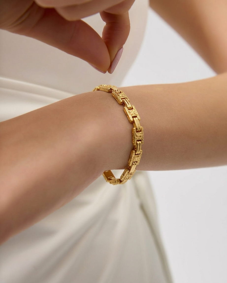 Persona abrochando una pulsera dorada de eslabones rectangulares en la muñeca, junto a una prenda blanca.