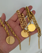 Tres pulseras doradas de cadenas de distintos grosores con discos redondos colgantes, sostenidas en una mano.