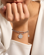 Persona con pulsera de tono plateado de eslabones y medalla redonda colgante, con la mano cerca del rostro y camisa blanca.