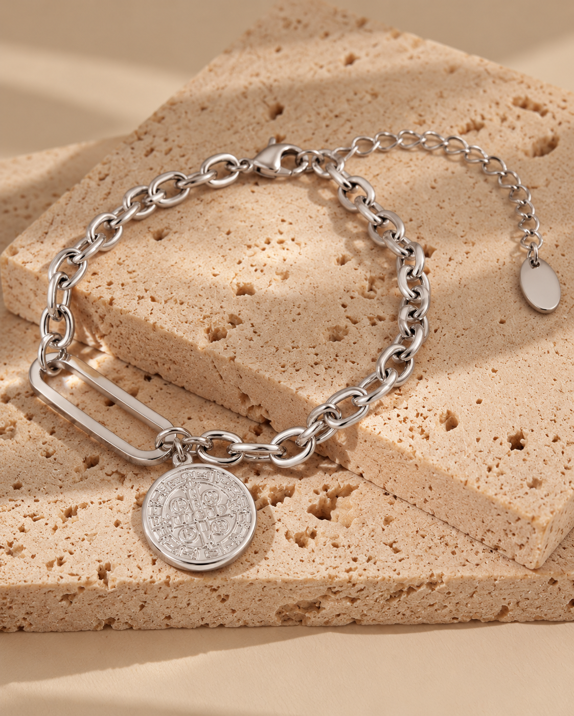 Pulsera de tono plateado con eslabones ovalados, eslabón central alargado y medalla redonda grabada, sobre piedra beige.