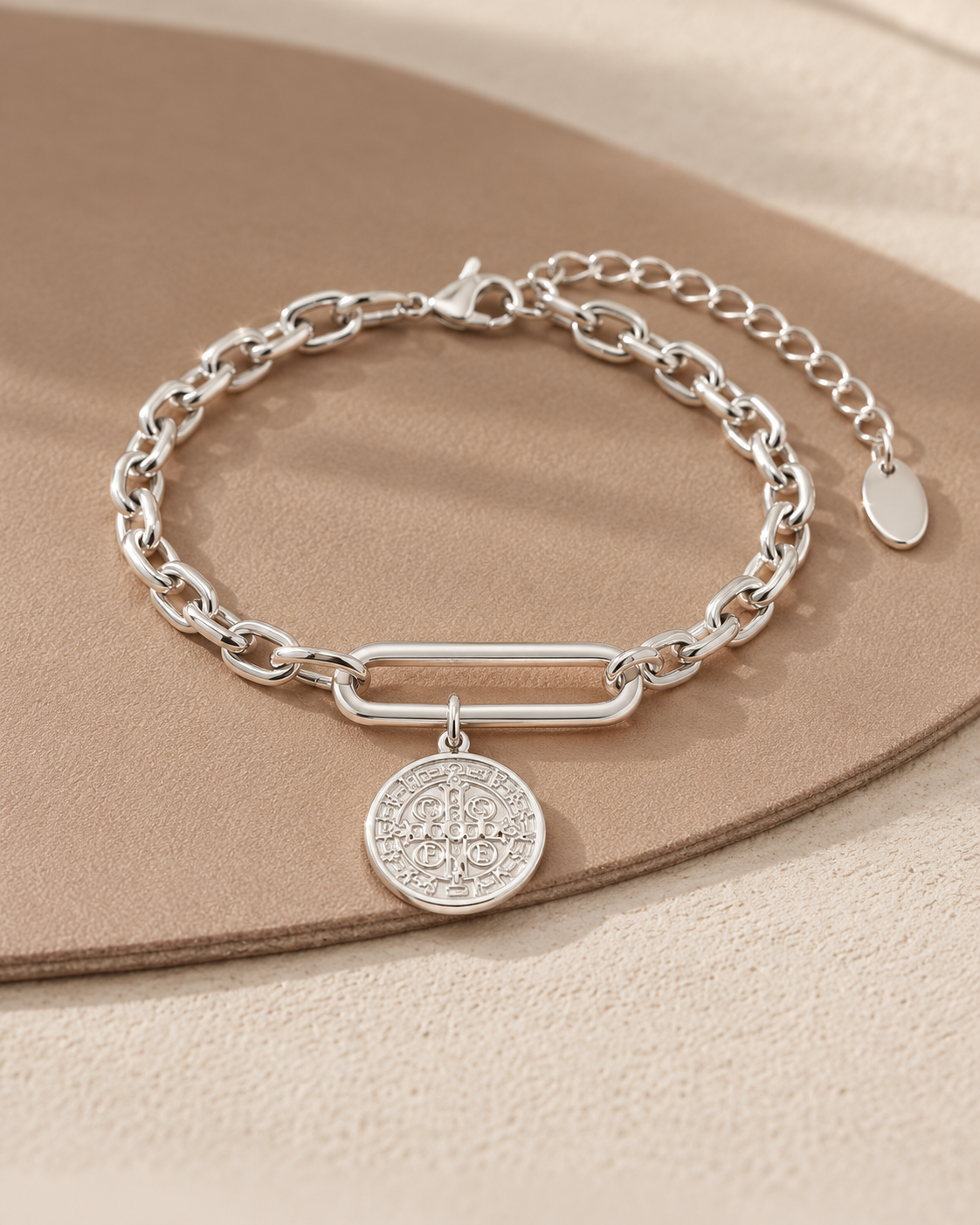 Pulsera de tono plateado con eslabón central alargado, medalla redonda grabada y cadena de extensión, sobre superficie beige.