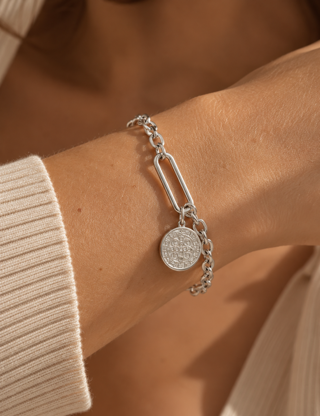Detalle de pulsera de tono plateado con eslabón alargado y medalla redonda grabada, puesta en la muñeca junto a manga de punto.