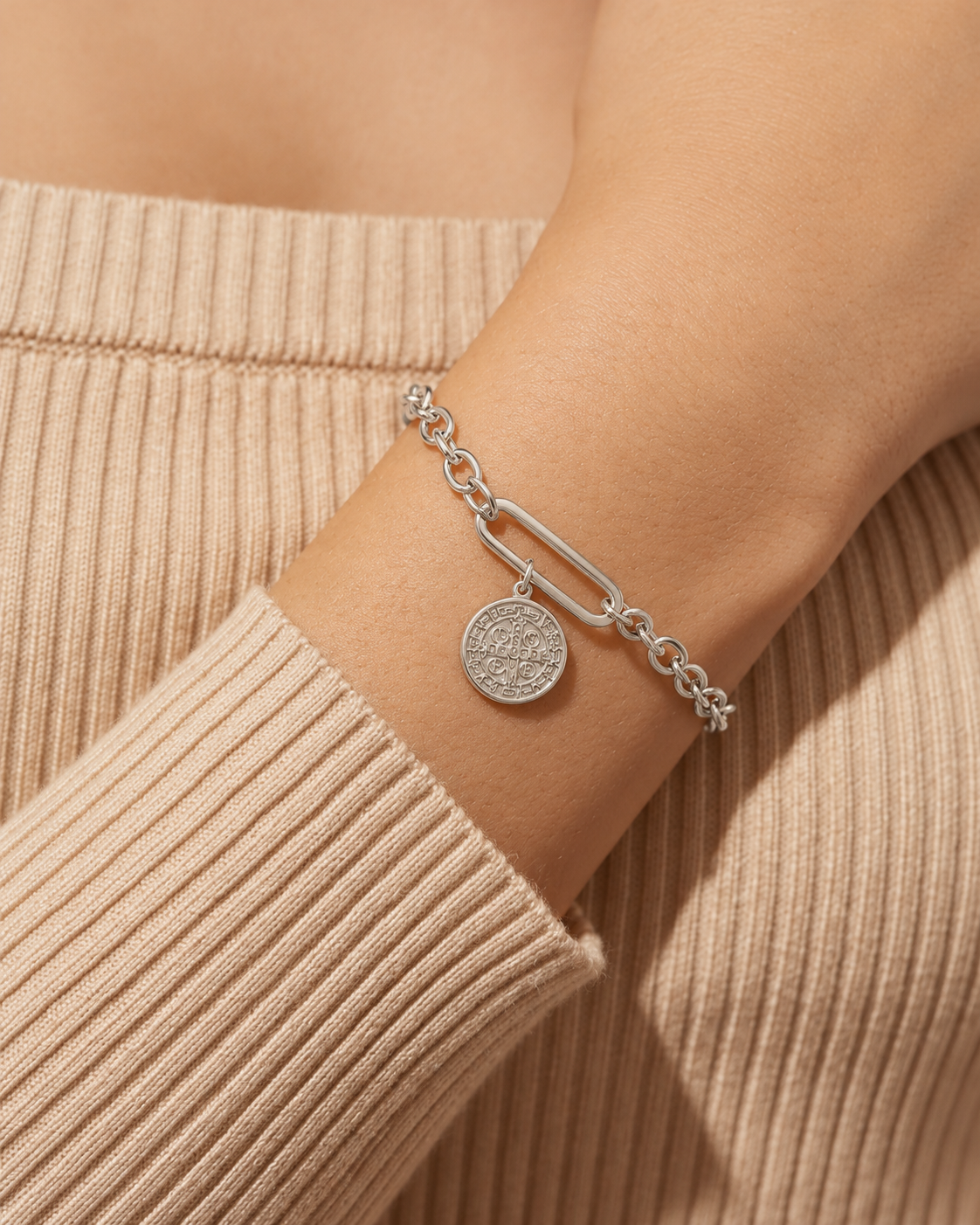 Pulsera de tono plateado de eslabones ovalados y uno alargado con medalla redonda colgante, puesta en la muñeca.