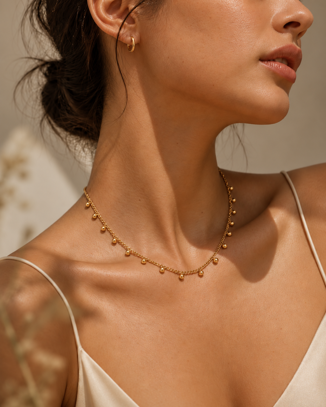 Mujer con collar fino dorado de pequeñas cuentas colgantes y pendientes de aro, con prenda clara de tirantes.