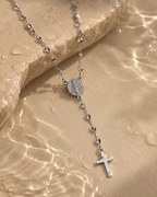Collar tipo rosario de tono plateado con medalla ovalada y cruz, sobre piedra junto al agua con reflejos de luz.